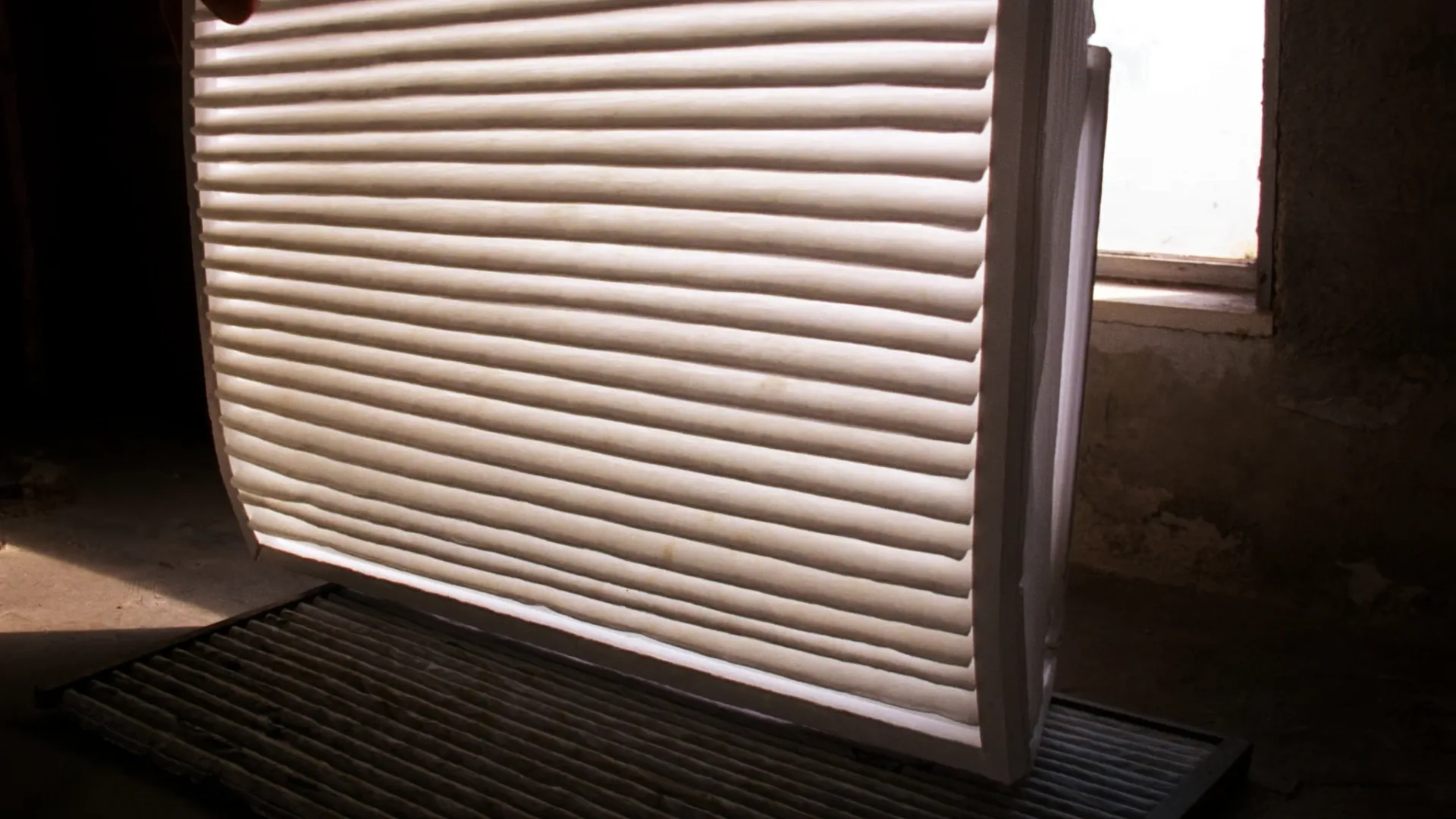 A clean pleated furnace filter held to the light beside a clogged grey one